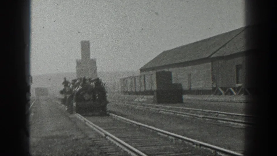 1936 California rail-yard view showing men packed on a small open rail car near tracks, a long building, and a smokestack.
