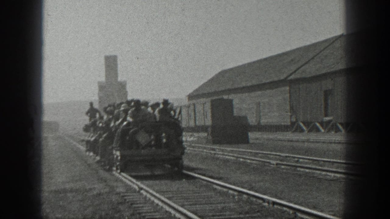 Men in hats crowd onto an open rail shuttle in a California rail yard, with rails, a long shed, and smokestack visible.