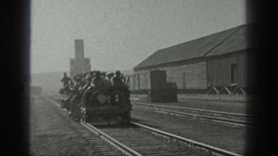 Men in hats crowd onto an open rail shuttle in a California rail yard, with rails, a long shed, and smokestack visible.