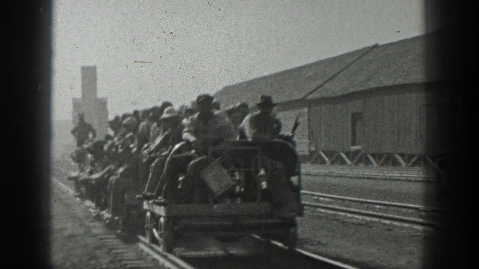 Rear view of a worker-filled open rail car in 1936 California, moving through a yard with tracks and a long building.