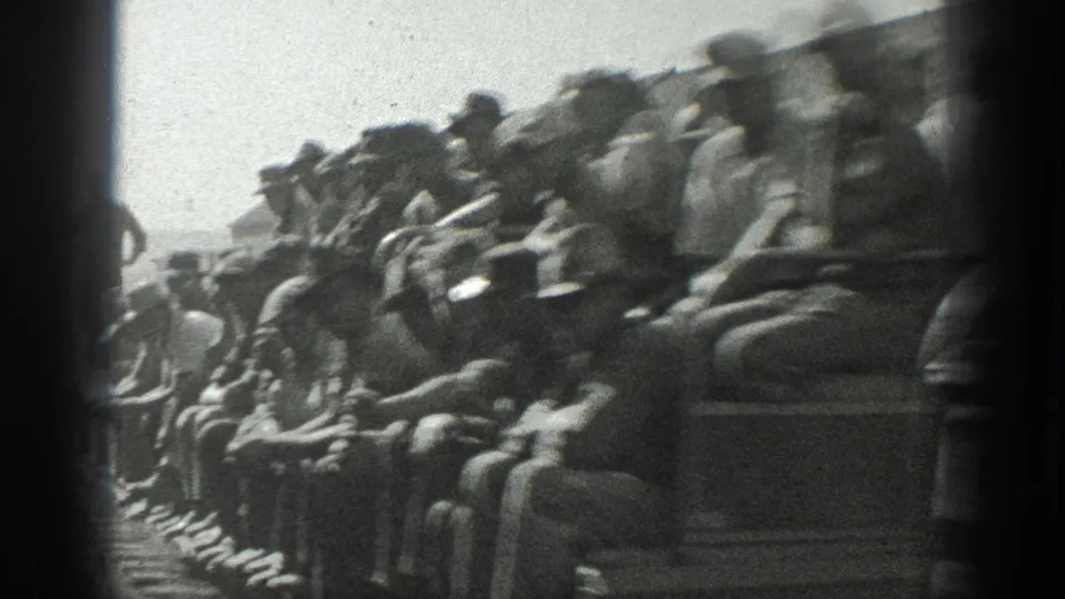 Close 8mm view of men in work clothes and hats crowded on an open rail shuttle in a California industrial yard.