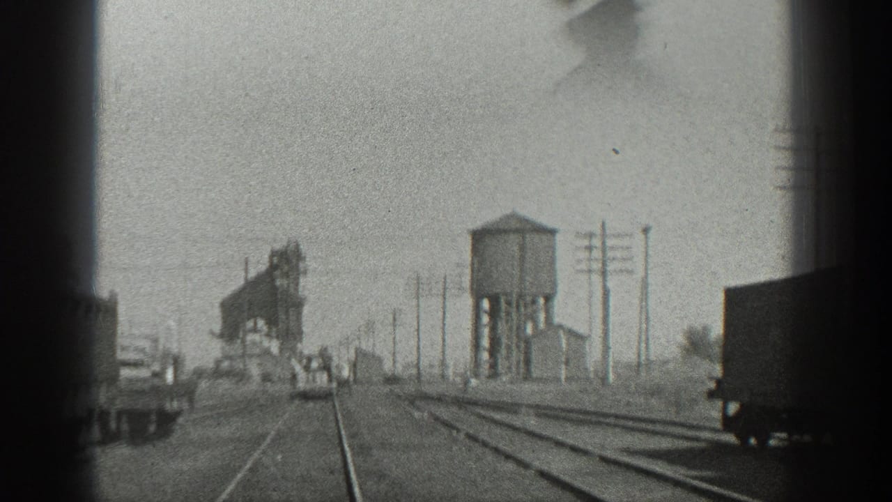 Rail tracks extend toward a water tower, freight cars, utility poles, and distant figures.