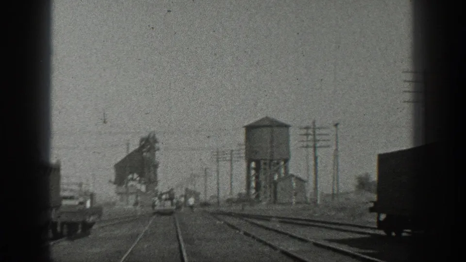 Black-and-white 8mm frame looking along California rail tracks toward a water tower and industrial rail structures.