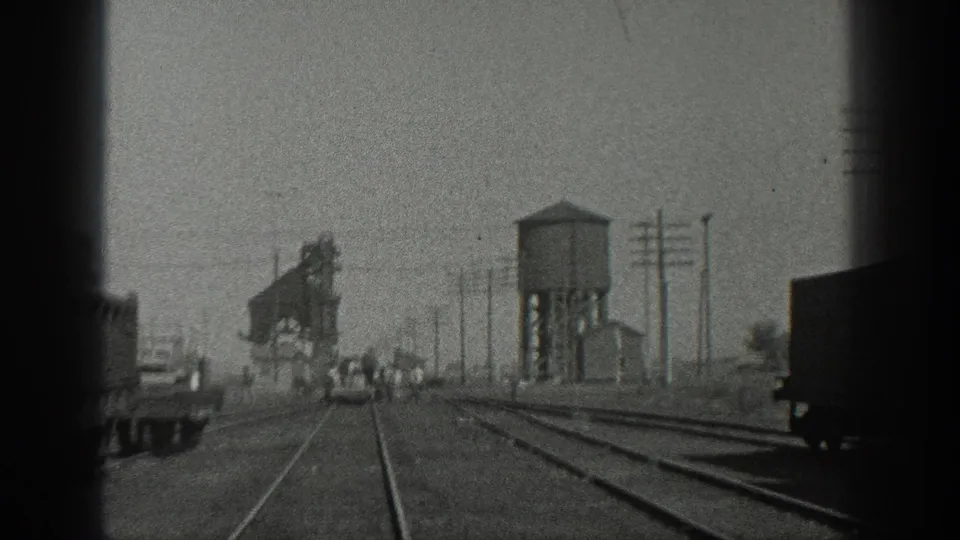 1936 California home-movie frame of a rail yard with tracks, freight cars, water tower, poles, and distant workers.
