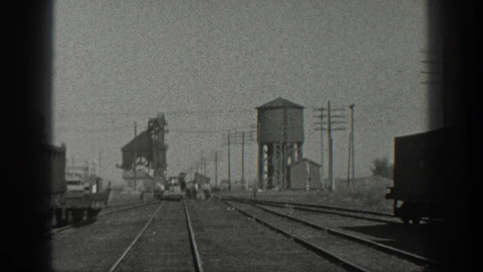 1936 California rail-yard view with converging tracks, distant people near a small rail car, utility poles, and a water tower-like structure.