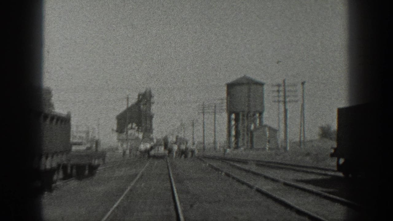 Rail tracks pass a dark rail car edge with people and a water tower farther ahead.