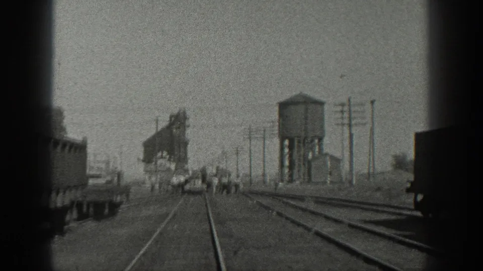 1936 California rail-yard frame showing a dark rail car at right, tracks leading to people near equipment, utility poles, and a water tower.