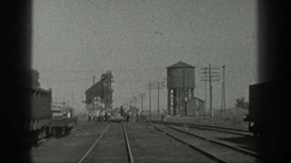 1936 California rail-yard scene with tracks converging toward distant people and rail equipment, framed by utility poles and a water tower-like structure.