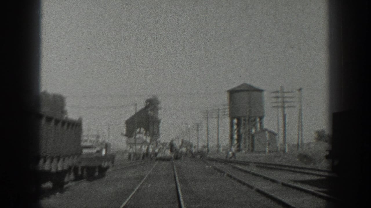 Railroad yard seen from a moving train with freight cars, tracks, water tower, and people in the distance.