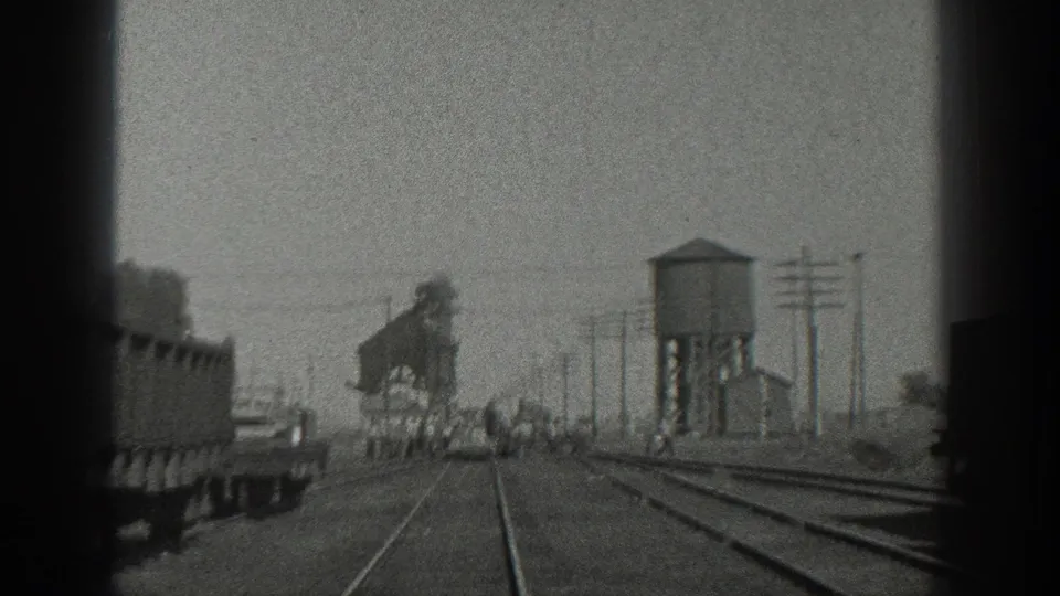 1936 California 8mm frame showing a railroad yard from a moving train, with parallel tracks, freight cars, a raised water tower, utility poles, and distant people.