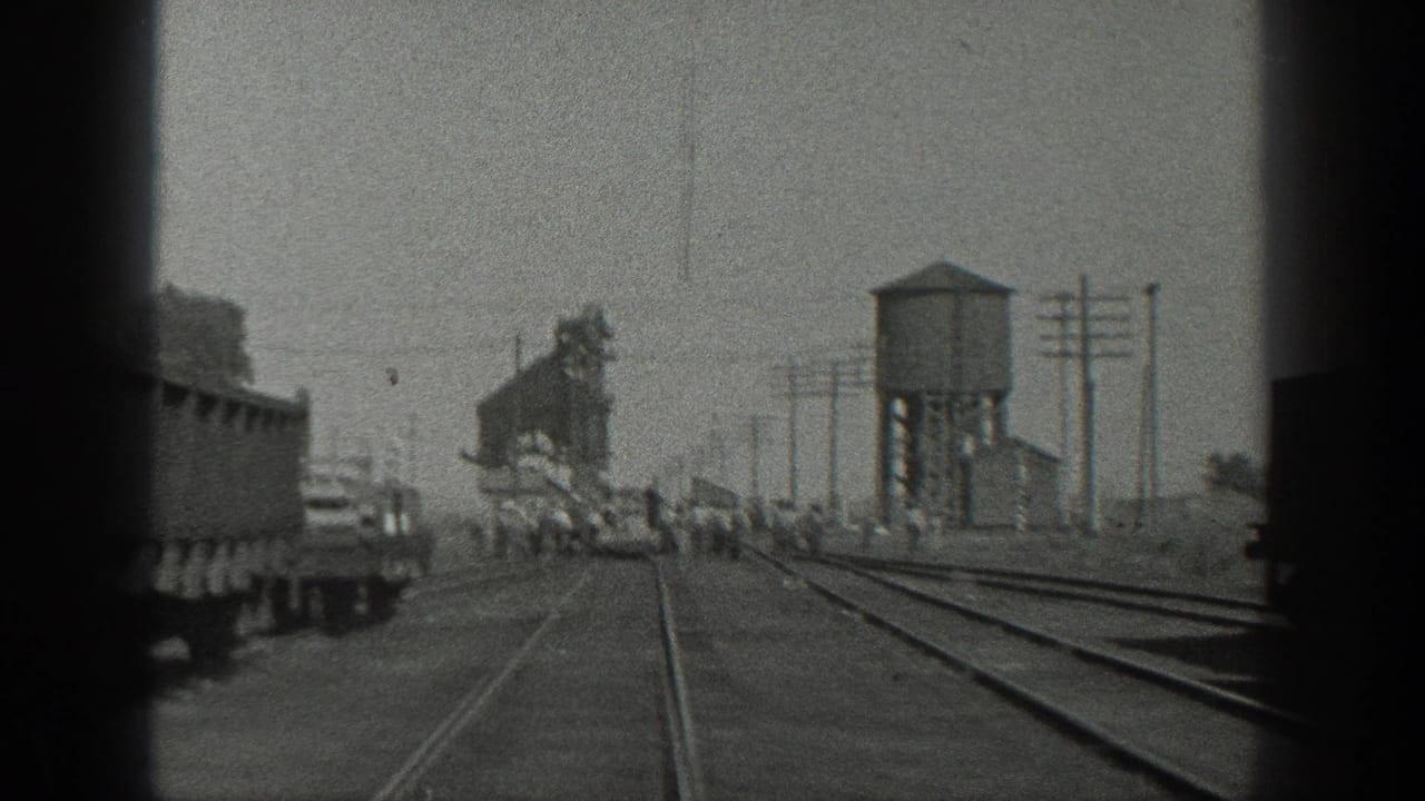 Railroad tracks run past freight cars and a water tower with people gathered near the rails.