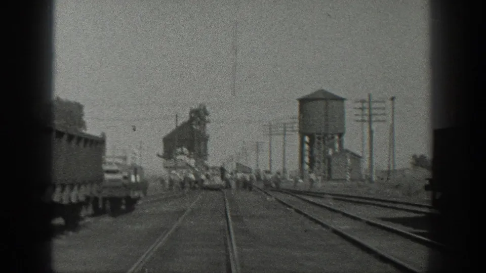 1936 California rail-yard view from a moving train, showing freight cars, converging tracks, a water tower, utility poles, and a small group of people near the track area.