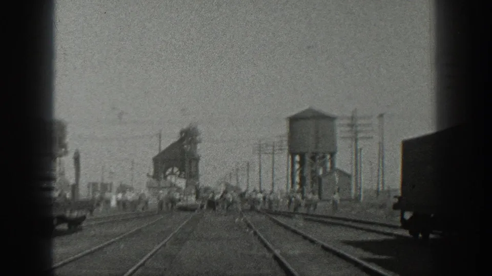 1936 California 8mm frame with a passing-train view across multiple tracks toward a rail-yard water tower, freight cars, utility poles, and people in the distance.