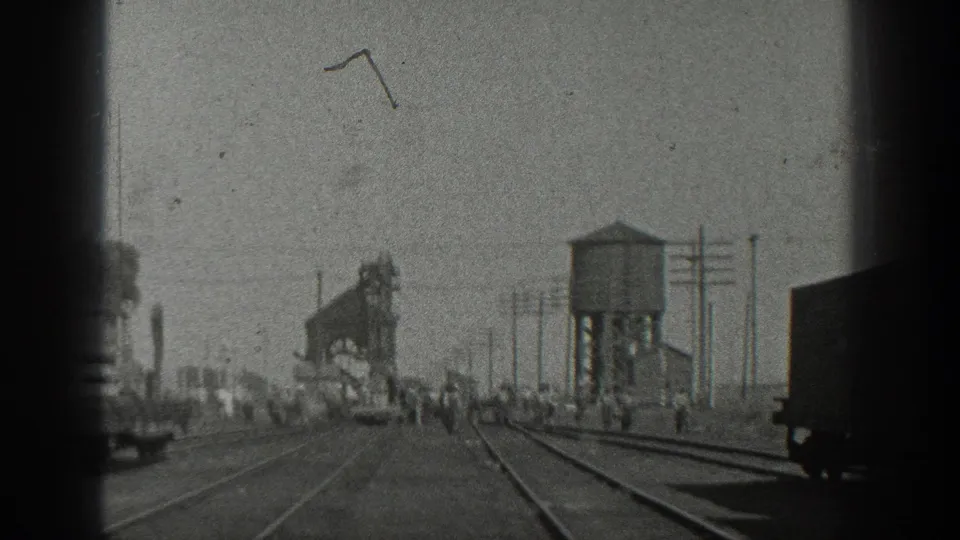 1936 California home-movie frame showing a railroad yard from a moving train, with dark frame edges, a water tower, utility poles, tracks, freight cars, and distant people.
