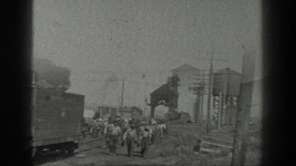 1936 California home-movie frame showing workers walking through a rail yard with freight cars, tracks, utility poles, and industrial structures.
