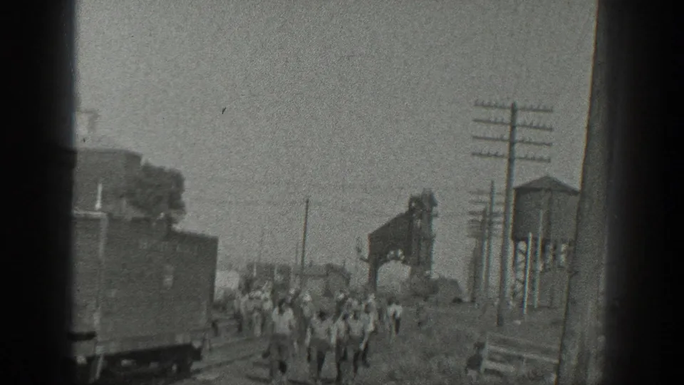 1936 California frame with workers walking through a rail yard beside tracks, a freight car, utility poles, and industrial equipment.