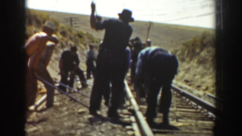 1936 California home-movie frame showing railroad workers doing track labor, with tools, rails, utility poles, and hillside terrain visible.
