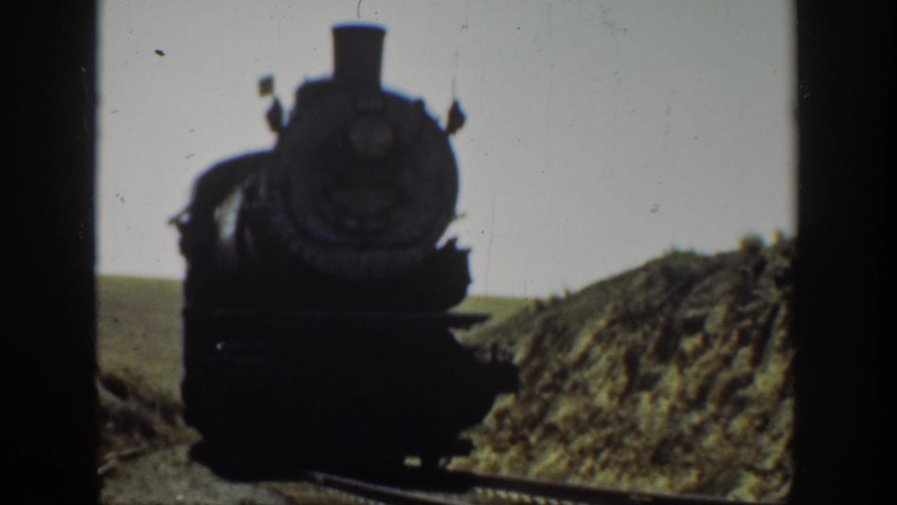 Steam locomotive closer to camera on track bordered by dirt banks.
