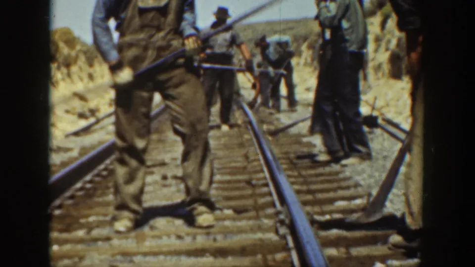 Several railroad workers maintain track with hand tools beside earth embankments in archival 8mm footage from California.