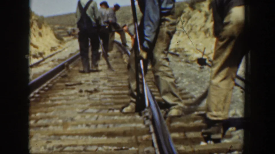 1936 California frame showing men gathered on a railroad track with tools, rails, ties, and dry hills visible beyond.
