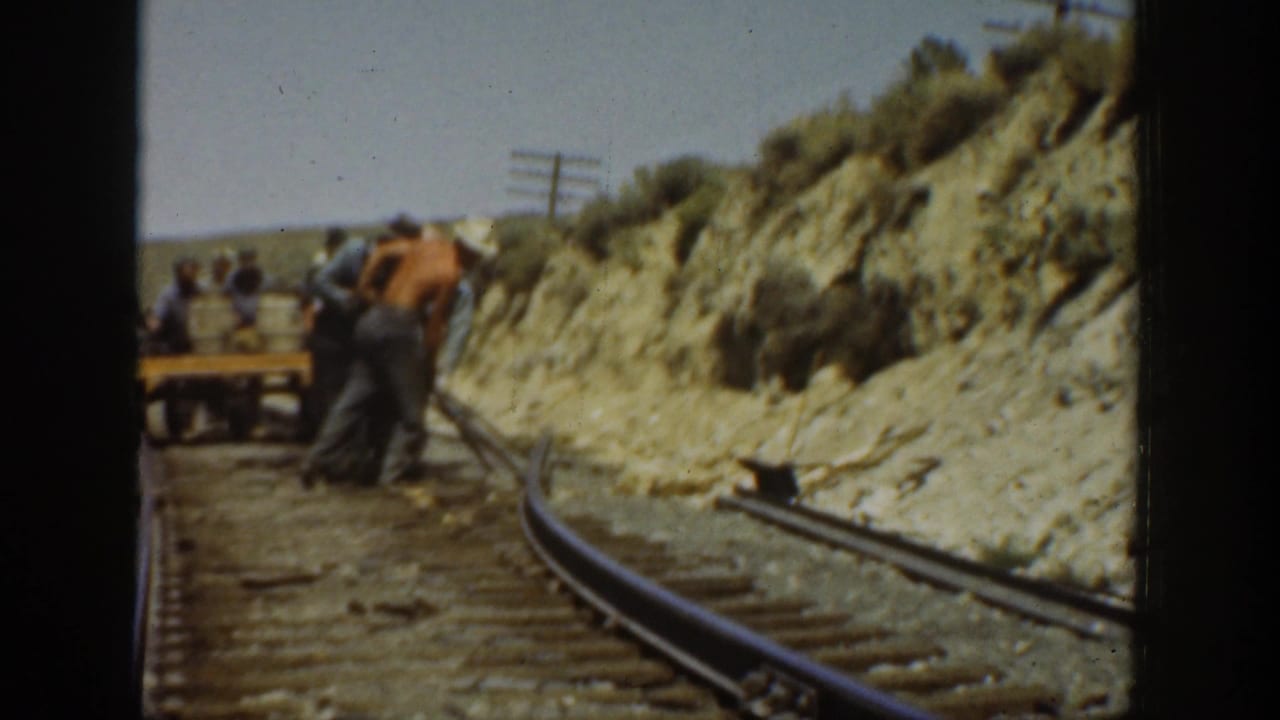 Men in work clothes adjust railway track in an arid California landscape, with rails curving past a cut bank.