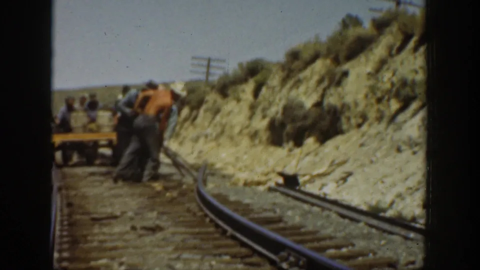 Men in work clothes adjust railway track in an arid California landscape, with rails curving past a cut bank.