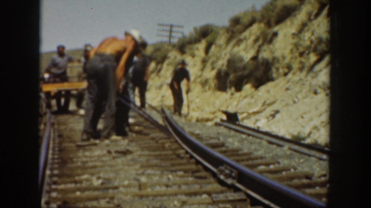 1936 California 8mm frame showing adult men using tools on a railroad track beside a sunlit dirt hillside.