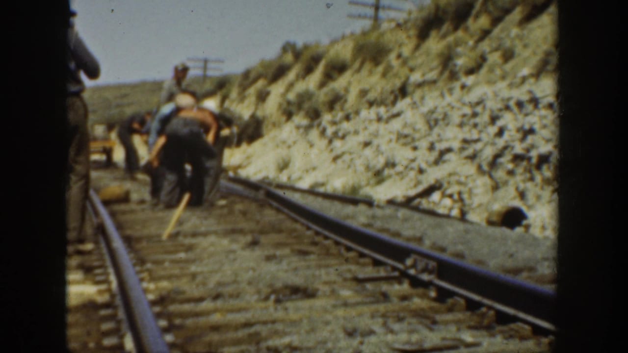 Workers in period clothing lean over a railroad track in California, with rails, ties, hillside, and utility poles visible.