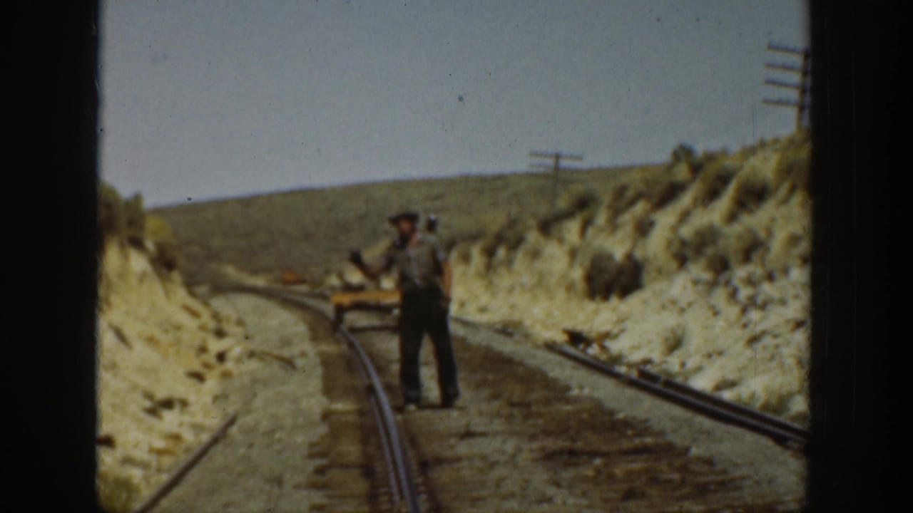 1936 California 8mm frame showing men near railroad tracks through a dry cut, with rails, hillside, and utility poles visible.