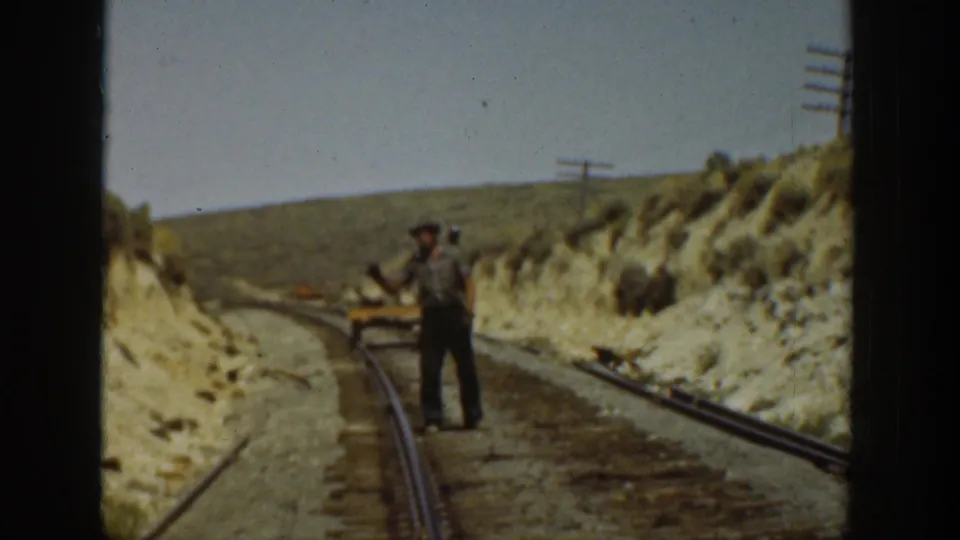 1936 California 8mm frame showing men near railroad tracks through a dry cut, with rails, hillside, and utility poles visible.