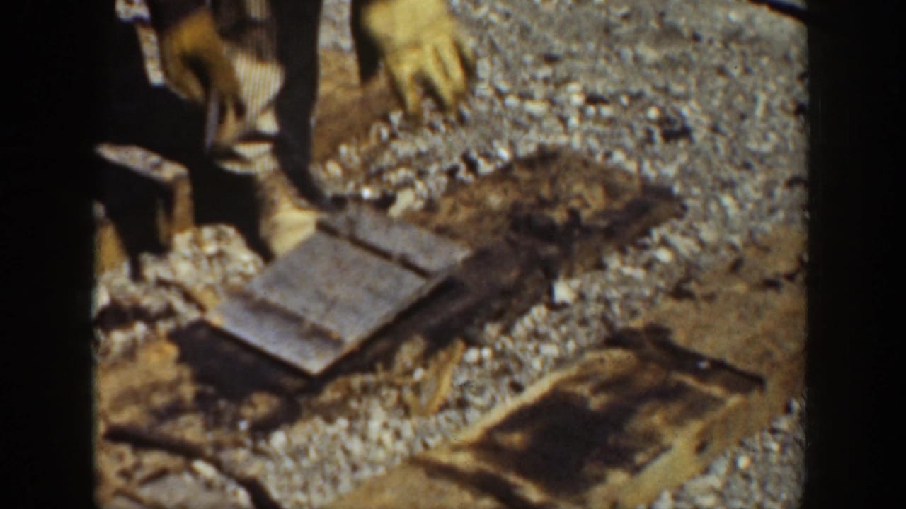 Close view of gloved hands and boots around wooden ties and tools on gravel ballast.