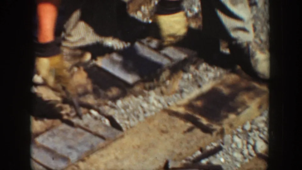 Close 8mm frame of railroad labor in California, with workers' legs, tools, timber ties, and ballast visible near the track.
