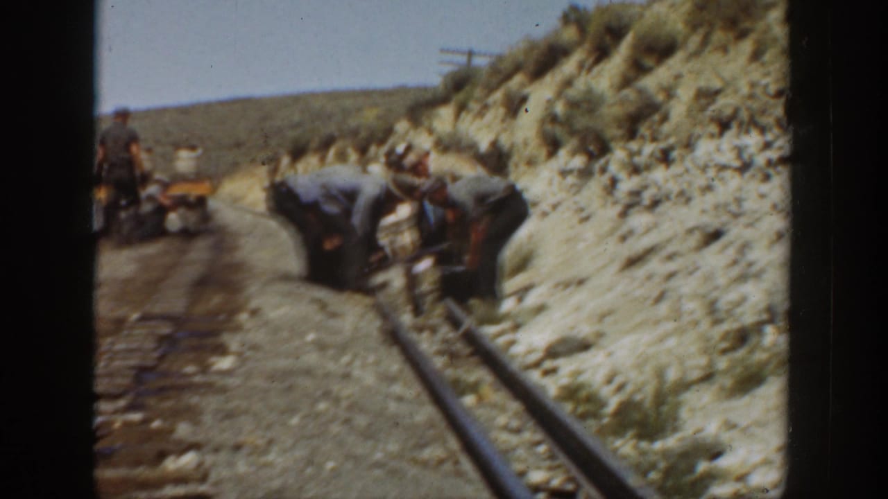 Men working beside railroad rails in a dry hillside cut with a small cart in the background.