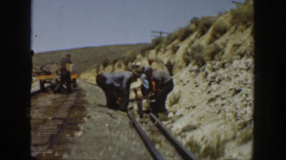 1936 California frame showing men clustered along a railroad track line, with a small cart and arid hillside terrain visible behind them.