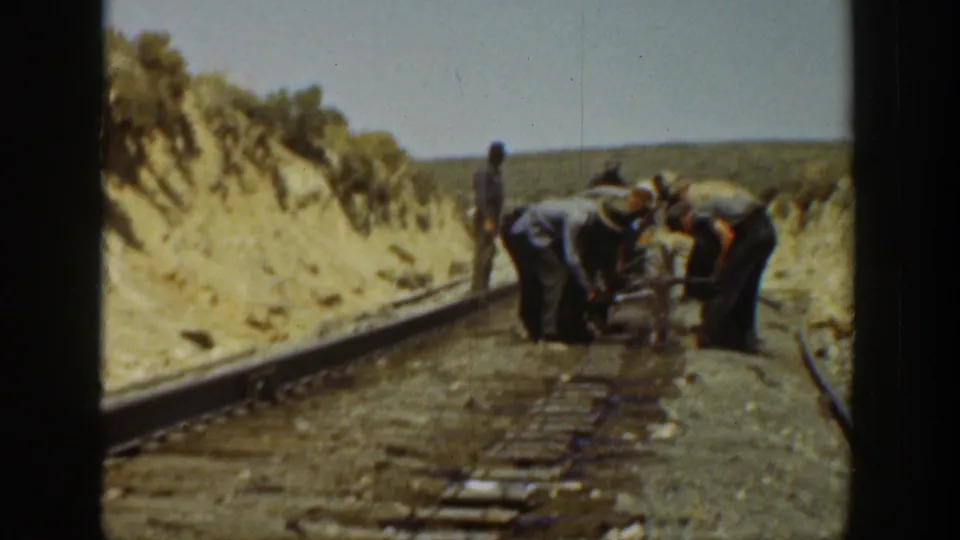 Workers in hats and work clothes lean over railroad track equipment in a dry California landscape, shown in soft 8mm color.