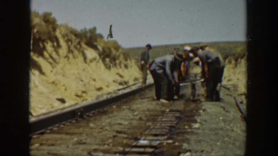 A fixed home-movie view shows men clustered at the center of railroad tracks in 1936 California, with dry banks and rails leading into the distance.