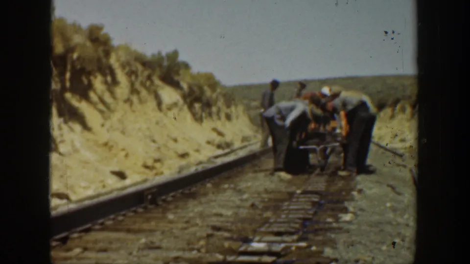 Men continue working on railroad tracks in a dry California rail cut, with hats, work clothes, rails, ties, and 8mm frame edges visible.