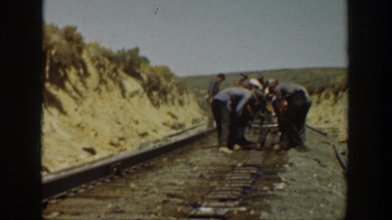 1936 California 8mm frame showing a small crew of men gathered over railroad rails in a dusty cut with scrub banks.
