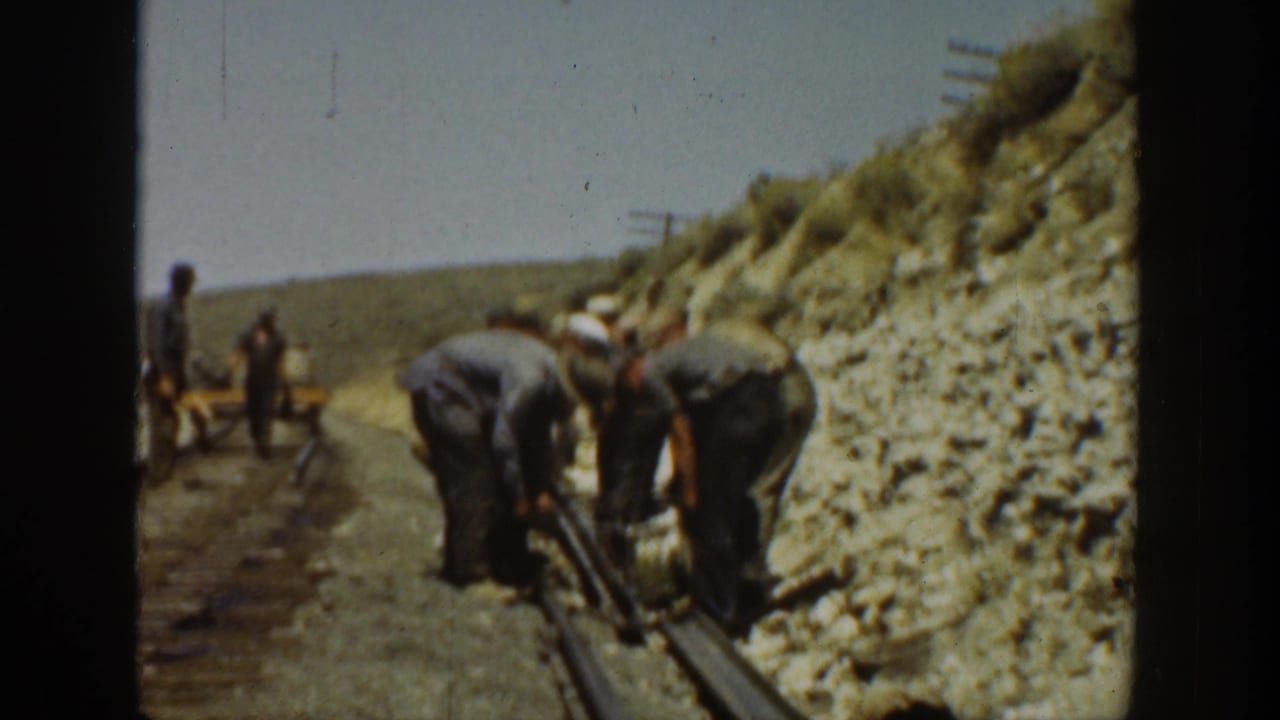 Workers bending over rail with tools on a dusty track bed.