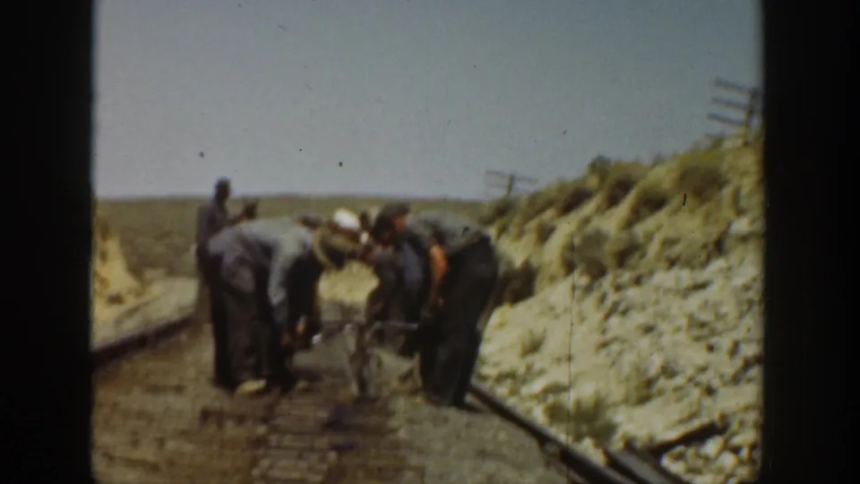 Workers lean close to the track, appearing to handle tools or rail hardware in a sunlit 1930s California railroad scene.