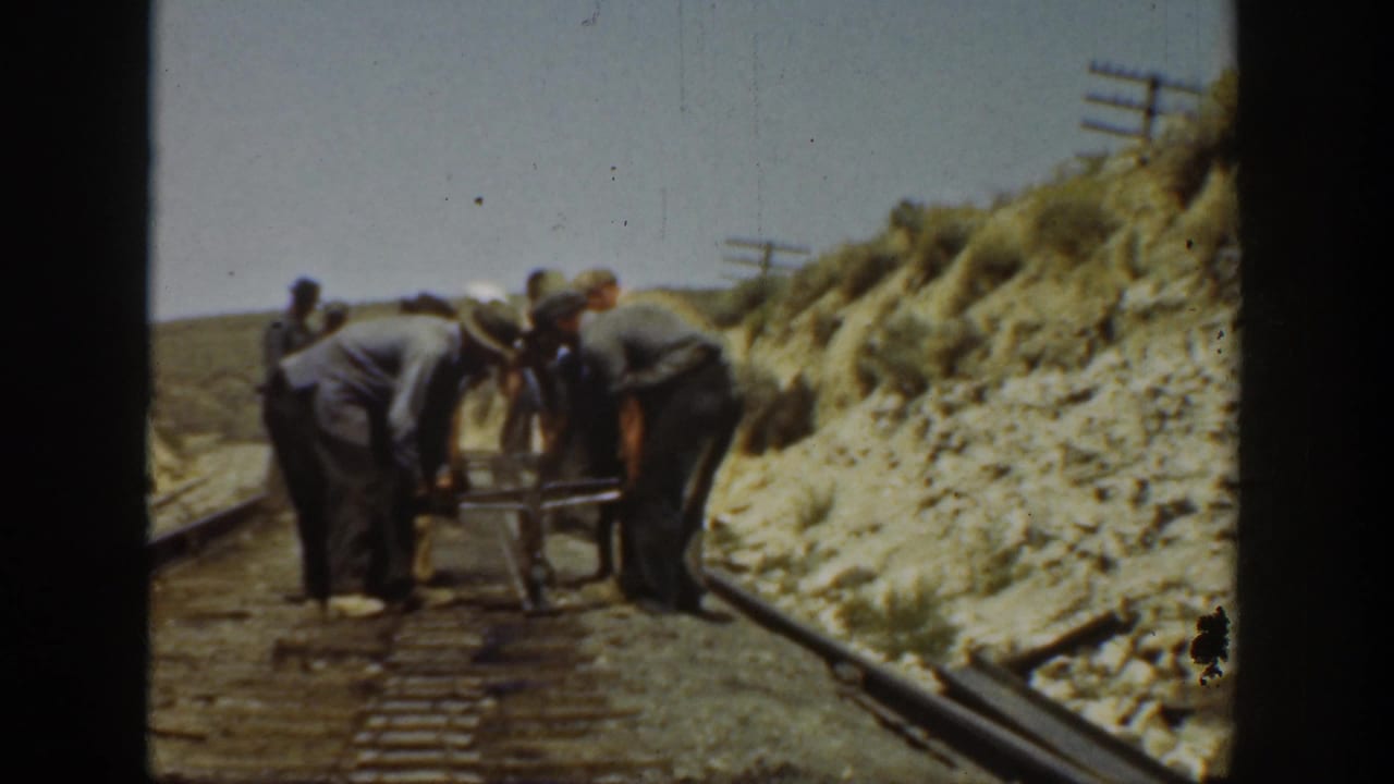 Men in work clothes bend over a rail tool on a track beside a dry embankment.
