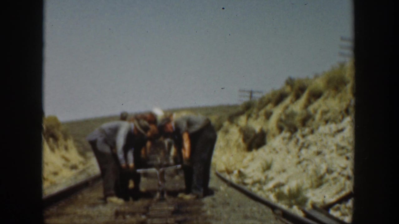 Adult workers appear to adjust or carry a heavy tool on a California rail line, with scrub-covered embankment behind them.