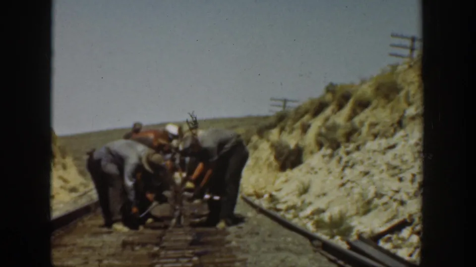 Several men in hats and work clothes bend over a rail tool between tracks in archival 1936 California home-movie footage.