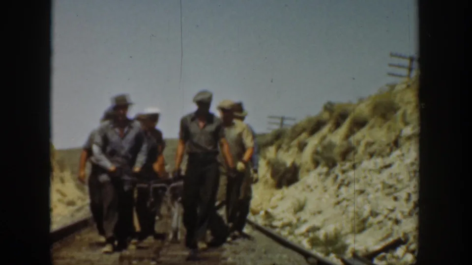 Three adult men in work clothes move toward the camera with a heavy rail tool on a dry California railroad track.