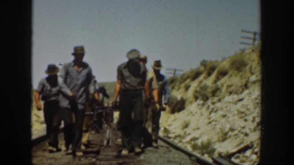1936 California 8mm frame showing adult men in work clothes walking along railway tracks in a bright, dry landscape.