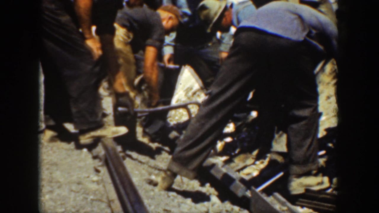Several men bend over railway track with tools and a wheelbarrow nearby.
