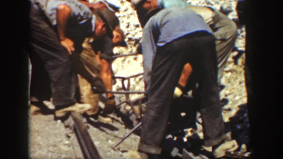 Close 8mm frame of men in work clothes bending beside a railway rail, with tools and gravel ballast visible.