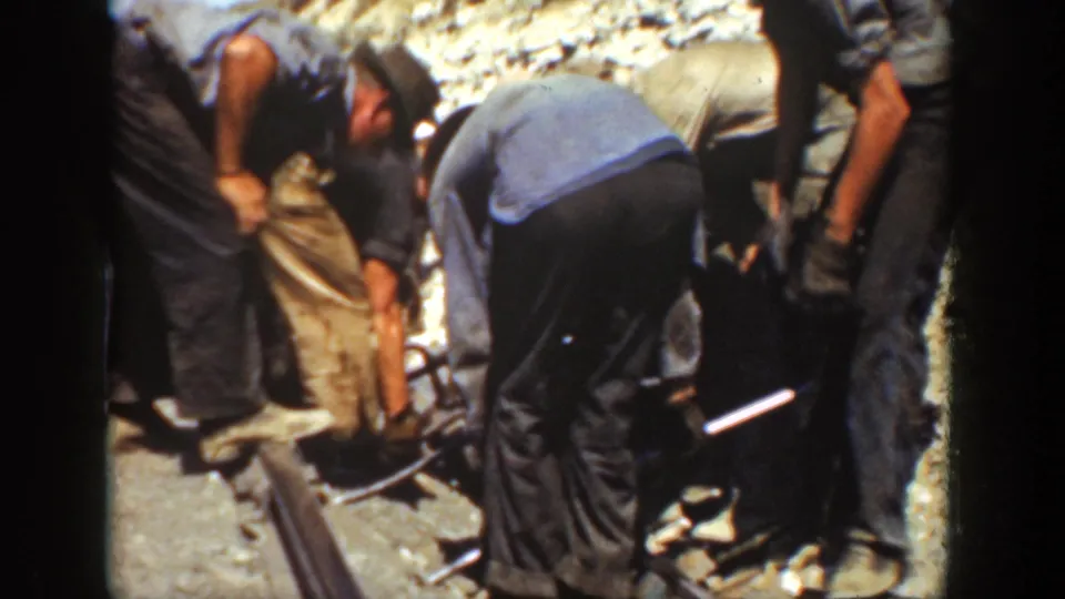 Adult men in work clothes bend over a railway track, with metal rails, tools, and sunlit ballast filling the frame.