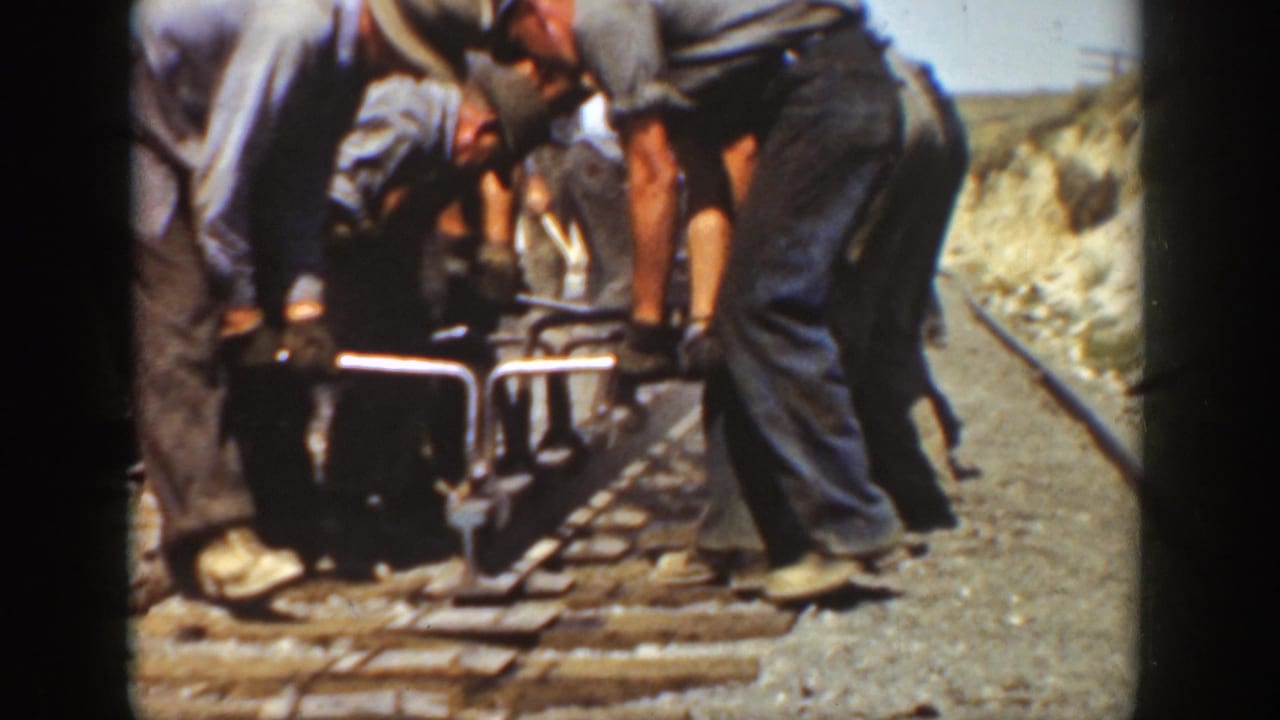 Adult men in work clothes appear to adjust or lay railroad track in 1936 California, with rails, wooden ties, and rocky ground visible.