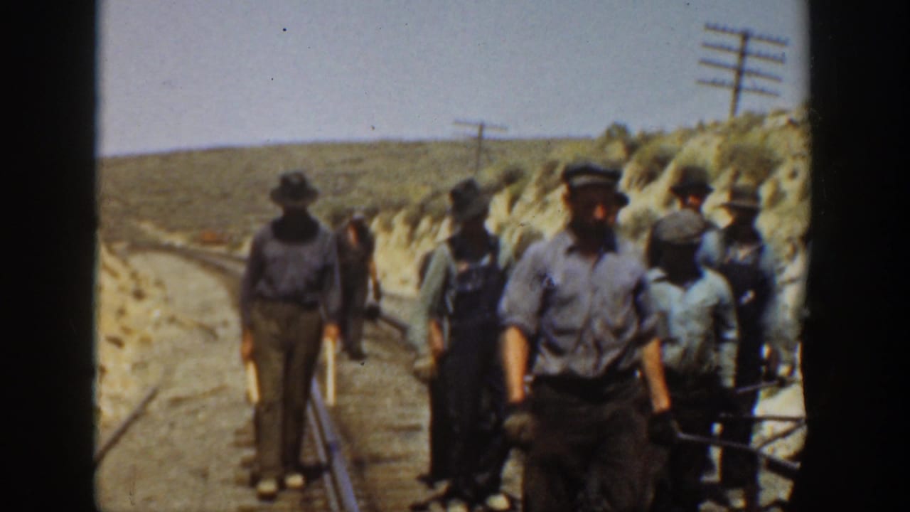 Railroad workers stand along a curved track bed in a dry California landscape, with rails, utility poles, hats, and work shirts visible.